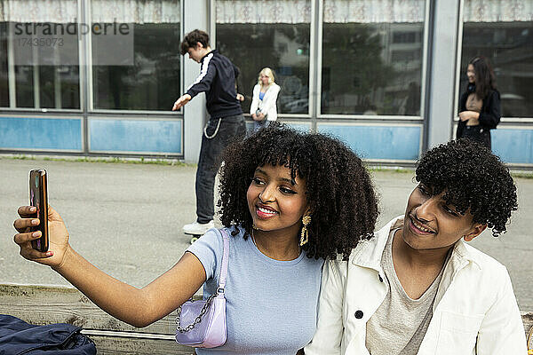 Teenager-Mädchen nimmt Selfie mit männlichen Freund durch Smartphone beim Sitzen auf Bank