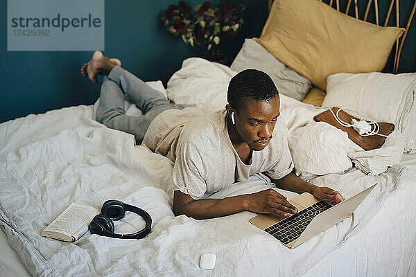 Mittlerer Erwachsener  der einen Laptop benutzt und dabei drahtlose In-Ear-Kopfhörer im Schlafzimmer trägt