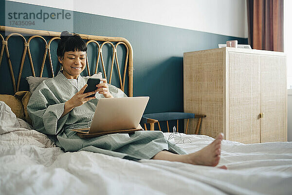 Lächelnde Frau mit Laptop und Smartphone auf dem Bett zu Hause