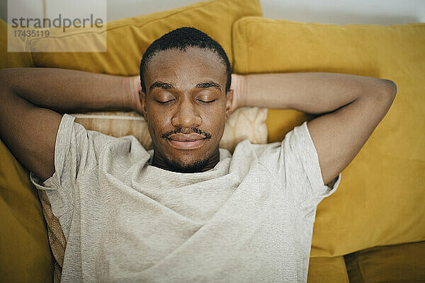 Mittlerer erwachsener Mann schläft mit den Händen hinter dem Kopf auf dem Sofa im Wohnzimmer