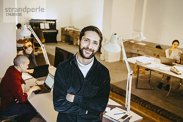 Porträt eines lächelnden männlichen Programmierers  der mit verschränkten Armen steht  während seine Kollegen am Schreibtisch im Büro arbeiten