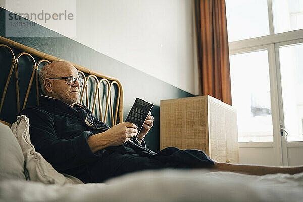 Älterer Mann liest ein Buch  während er auf dem Bett im Schlafzimmer sitzt