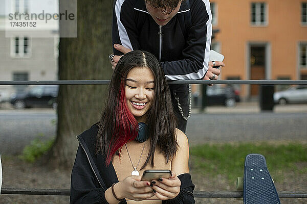 Teenager  der eine lächelnde Freundin betrachtet  die ein Smartphone im Park benutzt