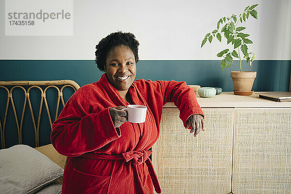 Porträt einer lächelnden erwachsenen Frau im Bademantel  die im Schlafzimmer Kaffee trinkt