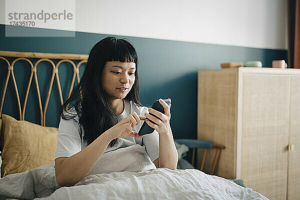 Junge Frau mit Smartphone auf dem Bett im Schlafzimmer