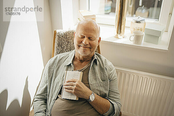 Lächelnder älterer Mann mit Buch  der sich auf einem Stuhl zu Hause entspannt