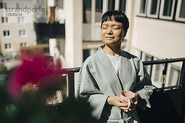 Lächelnde Frau hält eine Kaffeetasse und steht mit geschlossenen Augen auf einem Balkon