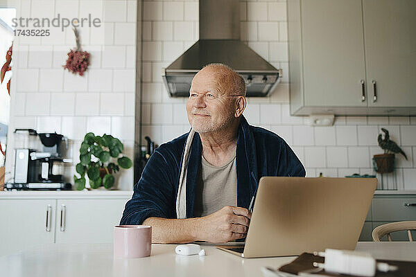 Lächelnder älterer Mann mit Laptop  der wegschaut  während er in der Küche zu Hause sitzt