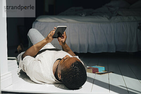 Mittelgroßer erwachsener Mann  der ein Mobiltelefon benutzt  während er zu Hause im Sonnenlicht auf dem Boden liegt