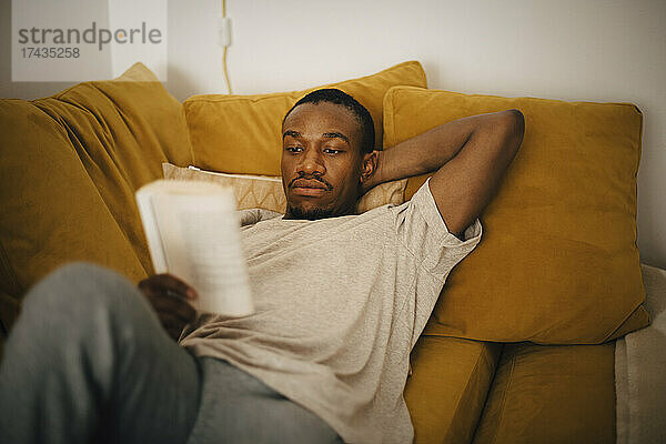 Mittlerer erwachsener Mann liest ein Buch  während er zu Hause auf dem Sofa liegt