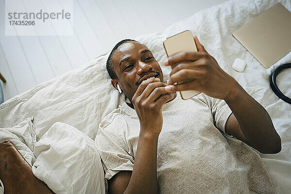 Lächelnder mittelgroßer erwachsener Mann  der ein Mobiltelefon benutzt  während er auf dem Bett liegt