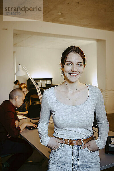 Porträt einer lächelnden Geschäftsfrau mit Händen in den Taschen  während ein Kollege am Schreibtisch im Büro arbeitet