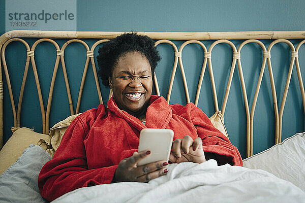 Glückliche Frau im mittleren Erwachsenenalter  die ihr Smartphone auf dem Bett zu Hause benutzt