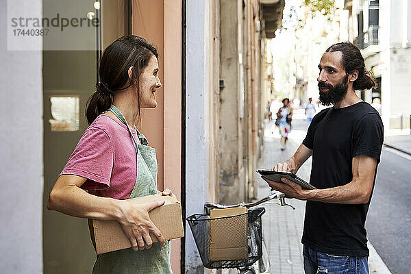 Männlicher Zusteller hält digitales Tablet im Gespräch mit Unternehmerin vor dem Geschäft