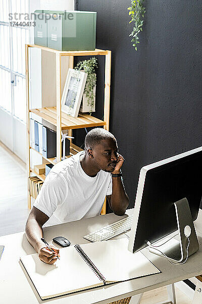 Männlicher Freiberufler sitzt mit Spiralnotizbuch und blickt im Studio auf den Desktop-Computer