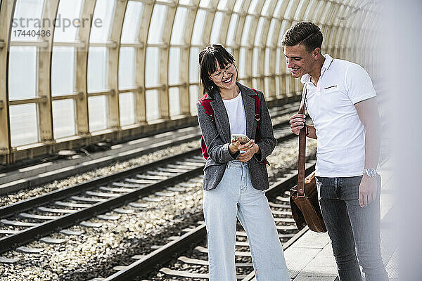 Glückliche männliche und weibliche Freunde teilen ihr Mobiltelefon am Bahnsteig