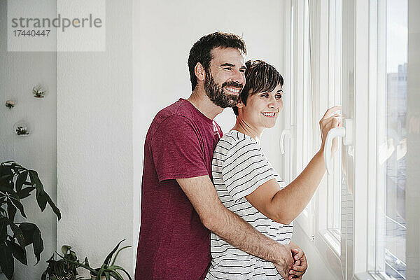 Mann umarmt Frau  während er zu Hause durch das Fenster schaut