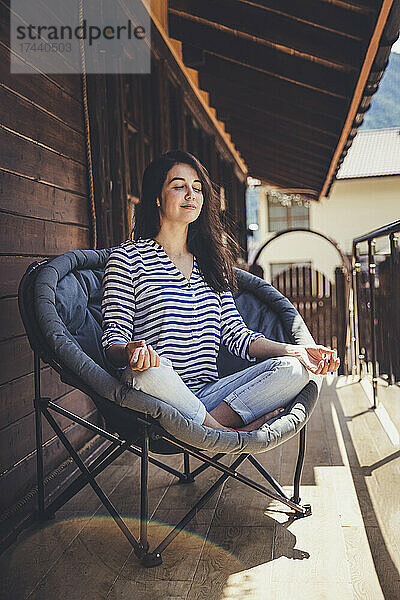 Frau meditiert  während sie mit gekreuzten Beinen auf einem Stuhl auf dem Balkon sitzt