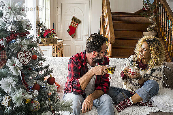 Lächelnde Frau trinkt Kaffee mit Mann  während sie zu Weihnachten zu Hause auf dem Sofa sitzt