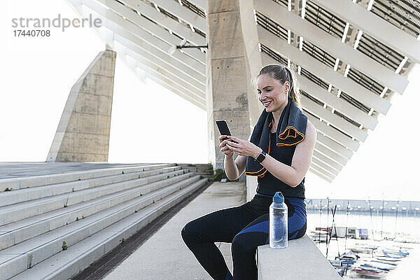 Lächelnde Sportlerin benutzt Smartphone  während sie auf Stützmauer sitzt