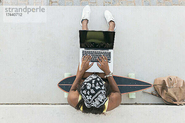 Woman wearing headscarf using laptop while sitting on skateboard