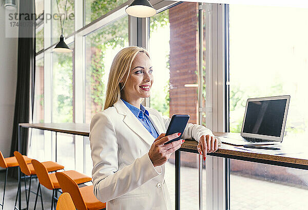 Geschäftsfrau mit Mobiltelefon und Laptop steht am Schreibtisch im Büro