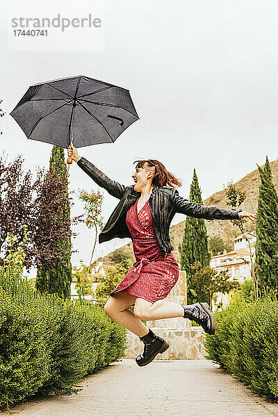 Glückliche Frau mittleren Alters springt bei Regen im Park mit Regenschirm