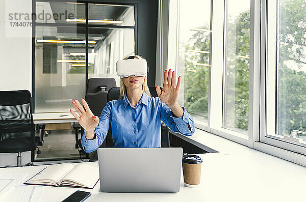 Geschäftsfrau gestikuliert  während sie im Büro ein Virtual-Reality-Headset trägt