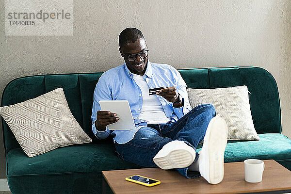 Lächelnder Mann mit digitalem Tablet  der auf die Kreditkarte schaut  während er zu Hause auf dem Sofa sitzt