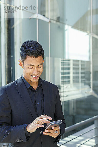 Lächelnder junger Geschäftsmann  der mit seinem Mobiltelefon im Internet surft  während er vor einem Bürogebäude steht