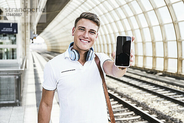 Lächelnder Mann zeigt Smartphone am Bahnhof