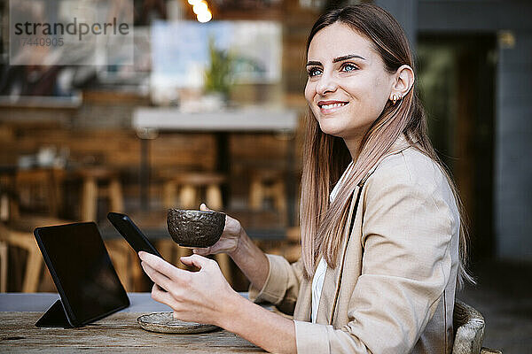 Lächelnde Geschäftsfrau mit Kaffeetasse und Smartphone auf der Caféterrasse