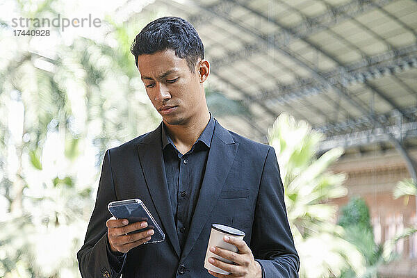 Männlicher Profi hält Einwegkaffeetasse in der Hand  während er sein Smartphone benutzt