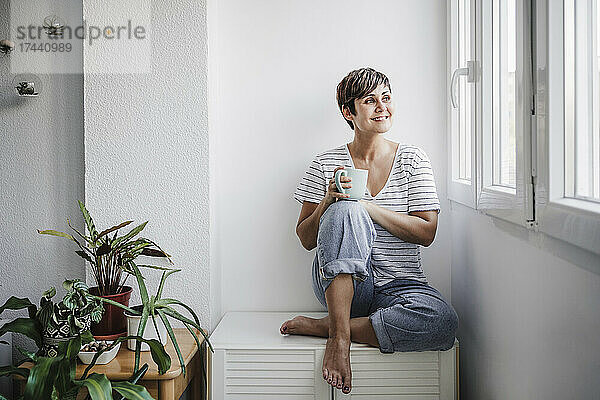 Lächelnde Frau mittleren Alters mit Tasse  die durch das Fenster schaut  während sie zu Hause sitzt