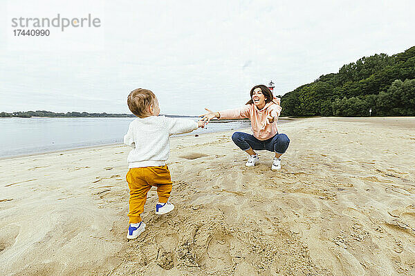 Männliches Kleinkind geht auf die Mutter zu  die am Strand im Sand kauert