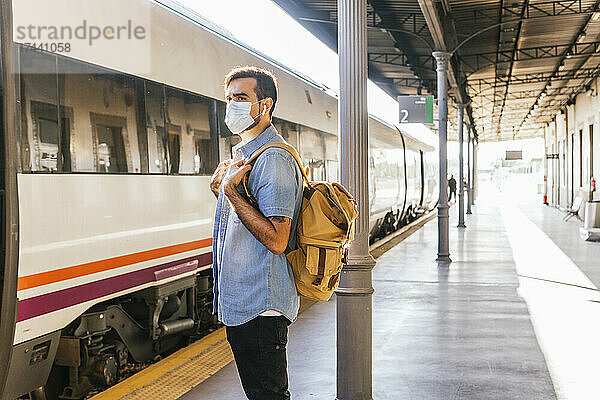 Junger Mann mit Rucksack steht während einer Pandemie auf dem Bahnsteig