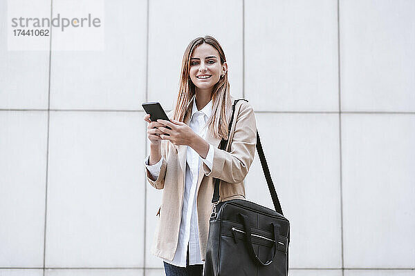 Lächelnde Geschäftsfrau mit Handy und Umhängetasche vor weißer Wand