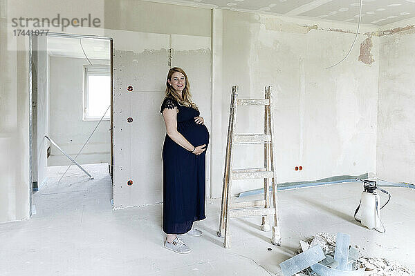 Lächelnde schwangere Frau berührt Bauch im Haus  das gerade renoviert wird
