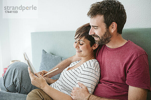 Ein Mann mittleren Alters schaut einer Frau zu  die in ein Notizbuch schreibt  während er zu Hause zusammen im Bett sitzt