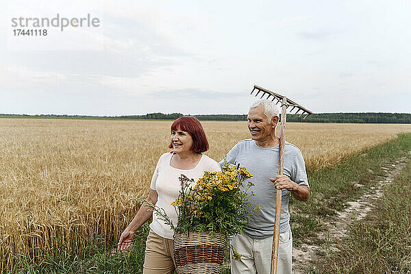 Älteres Paar mit Rainfarn-Blumenkorb und Rechen  das am Weizen vorbeigeht