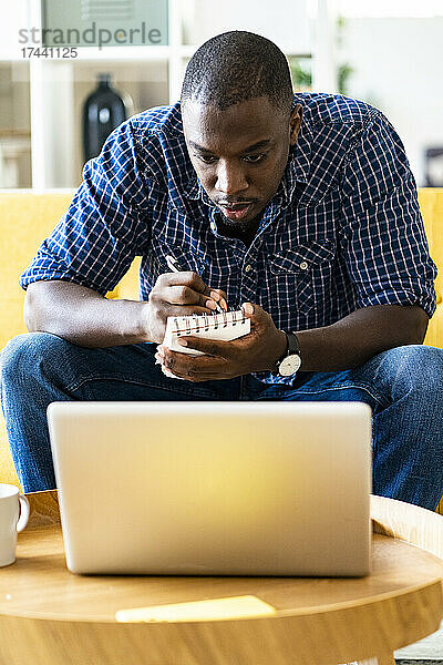 Mann schreibt Notizen in Notizblock  während er auf den Laptop schaut