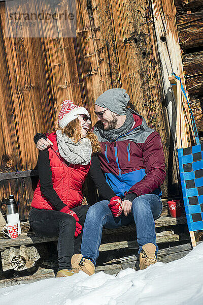 Mann mit Arm um Frau sitzt an sonnigen Wintertagen vor der Hütte