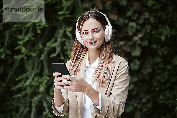 Geschäftsfrau hält Smartphone in der Hand und hört Musik über kabellose Kopfhörer