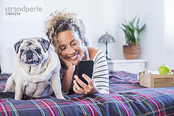 Lächelnde Frau benutzt Mobiltelefon  während sie mit Hund im Schlafzimmer auf dem Bett liegt