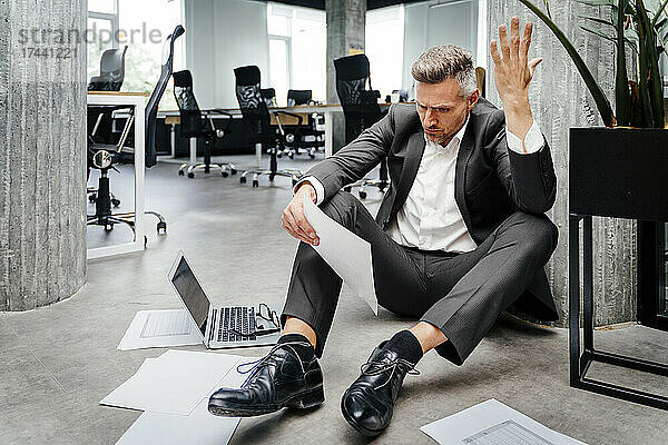 Frustrierter Geschäftsmann gestikuliert  während er im Büro ein Dokument betrachtet