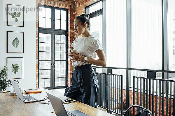 Geschäftsfrau mit Kaffeetasse und Blick auf den Laptop  während sie im Büro steht