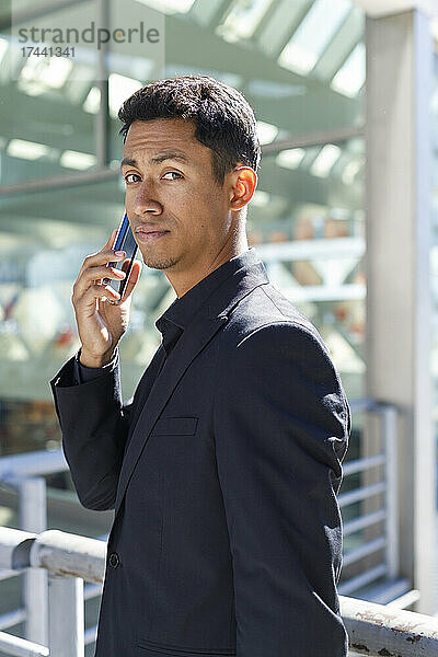 Hübscher männlicher Berufstätiger  der mit dem Mobiltelefon spricht  während er vor dem Bürogebäude steht