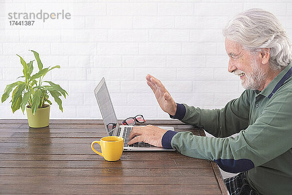 Lächelnder Mann winkt während eines Videoanrufs über den Laptop mit der Hand  während er am Tisch sitzt