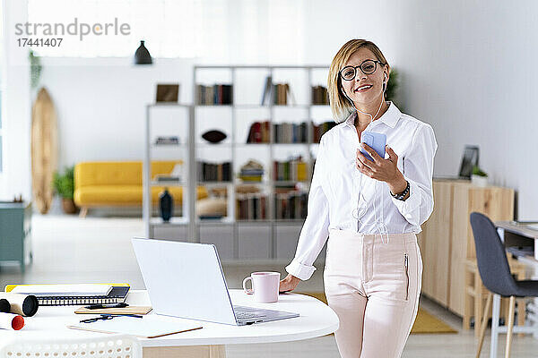 Lächelnde Berufstätige mit Brille  während sie ihr Smartphone am Schreibtisch hält