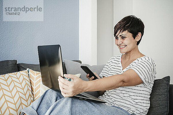 Lächelnde Freiberuflerin mit Smartphone und Laptop während eines Videoanrufs zu Hause